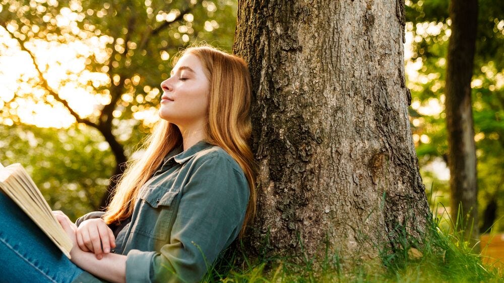Sieviete sēž zālē, atspiedusies pret koku un ar aizvērtām acīm bauda mierpilnu brīdi, lasot grāmatu zaļā, saulainā Dzirciema apkaimes ainavā